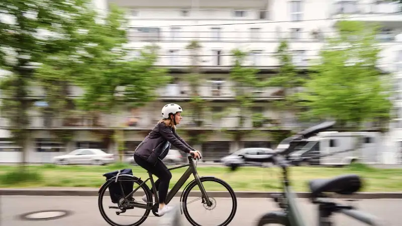 Eine Radfahrerin in Bürokleidung fährt die Straße entlang, Sie hat ihre Tasche am Hinterradblech ihres Fahrrads befestigt Eine Radfahrerin in Bürokleidung fährt die Straße entlang, Sie hat ihre Tasche am Hinterradblech ihres Fahrrads befestigt