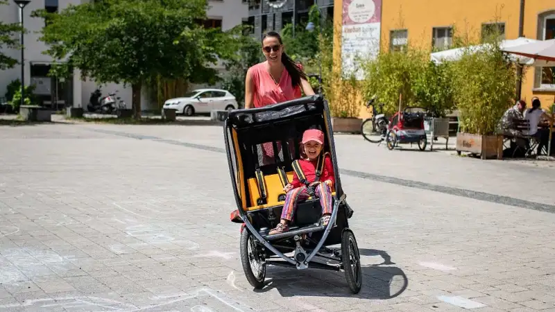 Eine Mutter fährt das Chike e-Bike in der Stadt. Ihr Kind sitzt angeschnallt darin. Beide lachen Eine Mutter fährt das Chike e-Bike in der Stadt. Ihr Kind sitzt angeschnallt darin. Beide lachen